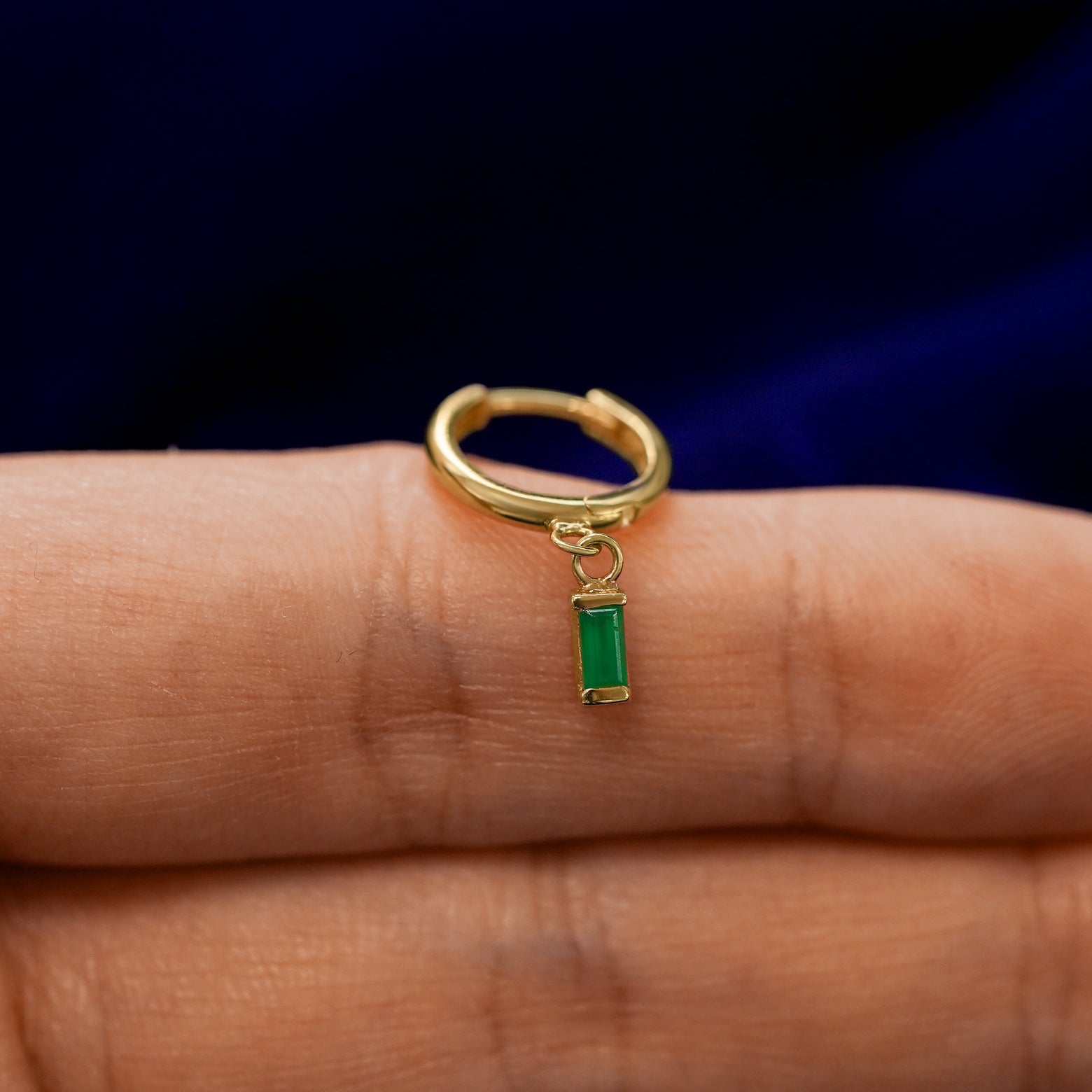 A small curvy huggie hoop earring with a dangling baguette green emerald charm balanced on a models finger