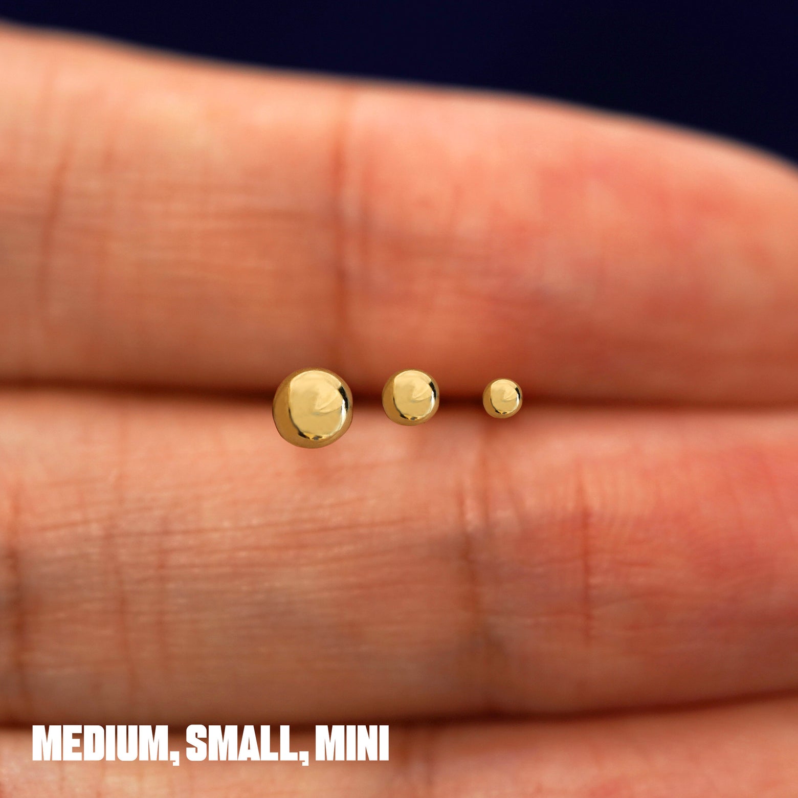 Solid 14k yellow gold Medium, Small, and Mini Ball Flatback Piercings in between a model's fingers