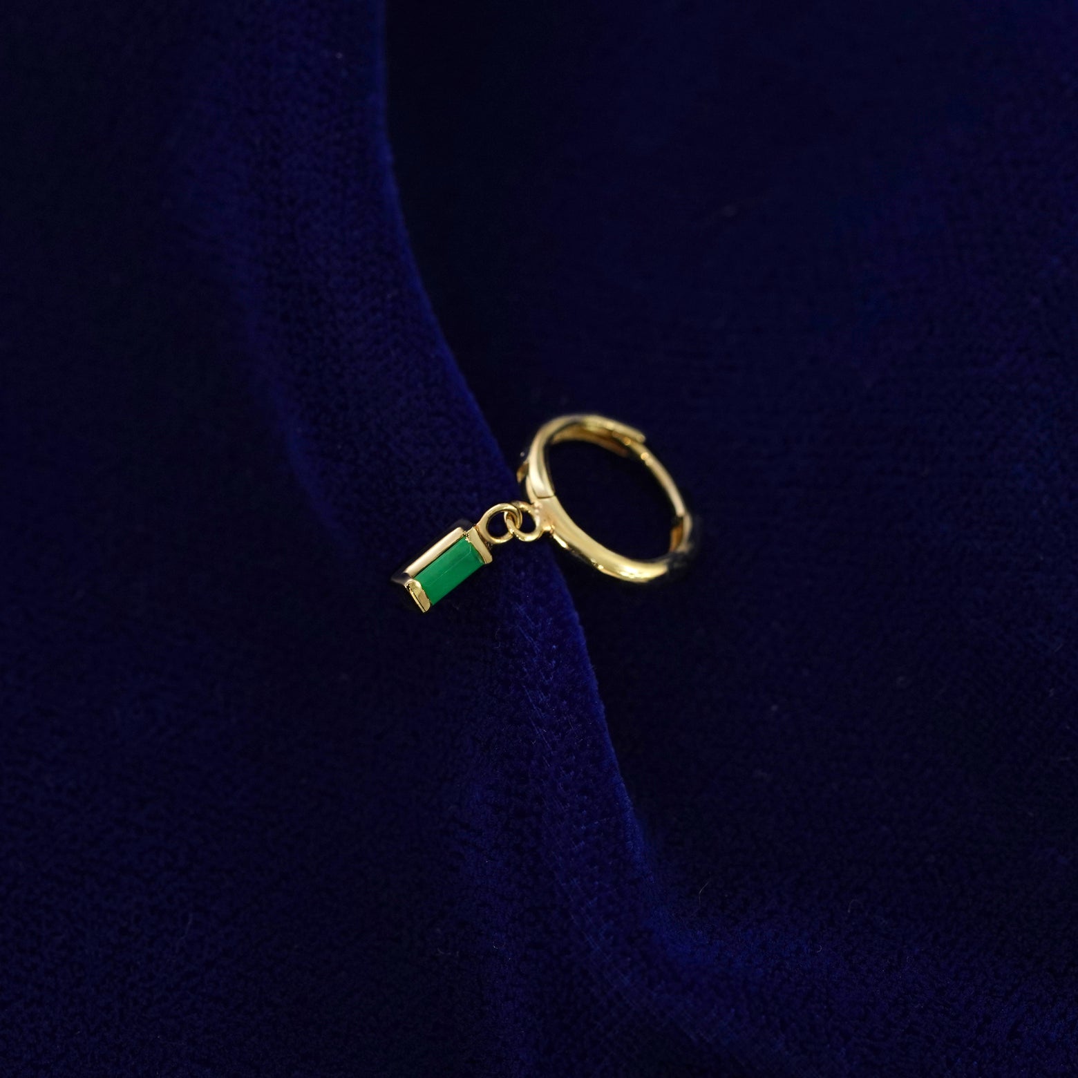 A recycled solid 14k gold Baguette Emerald Dangle Huggie Hoop earring on a dark blue background