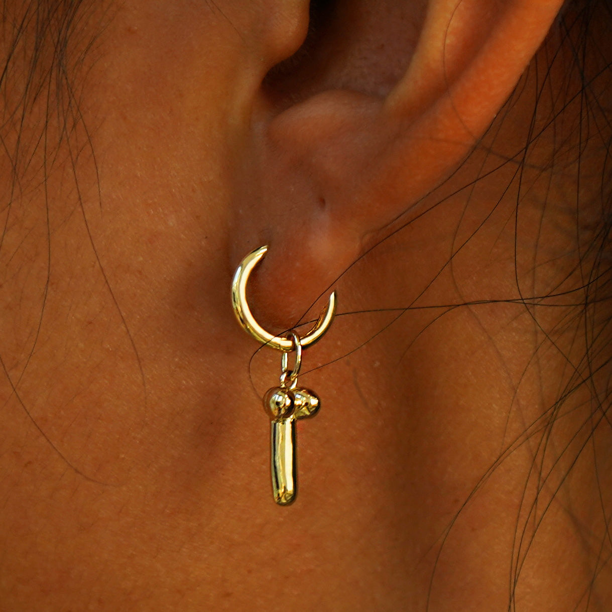 Close up view of a model's ear wearing a yellow gold Penis Charm on a Curvy Huggie Hoop