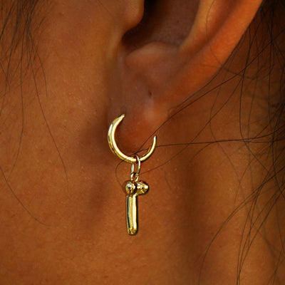 Close up view of a model's ear wearing a yellow gold Penis Charm on a Curvy Huggie Hoop