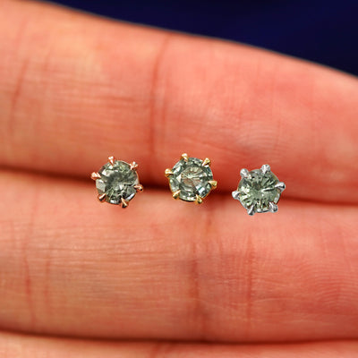 Three versions of the Montana Teal Sapphire Earring in options of yellow, white, and rose gold in between a model's fingers