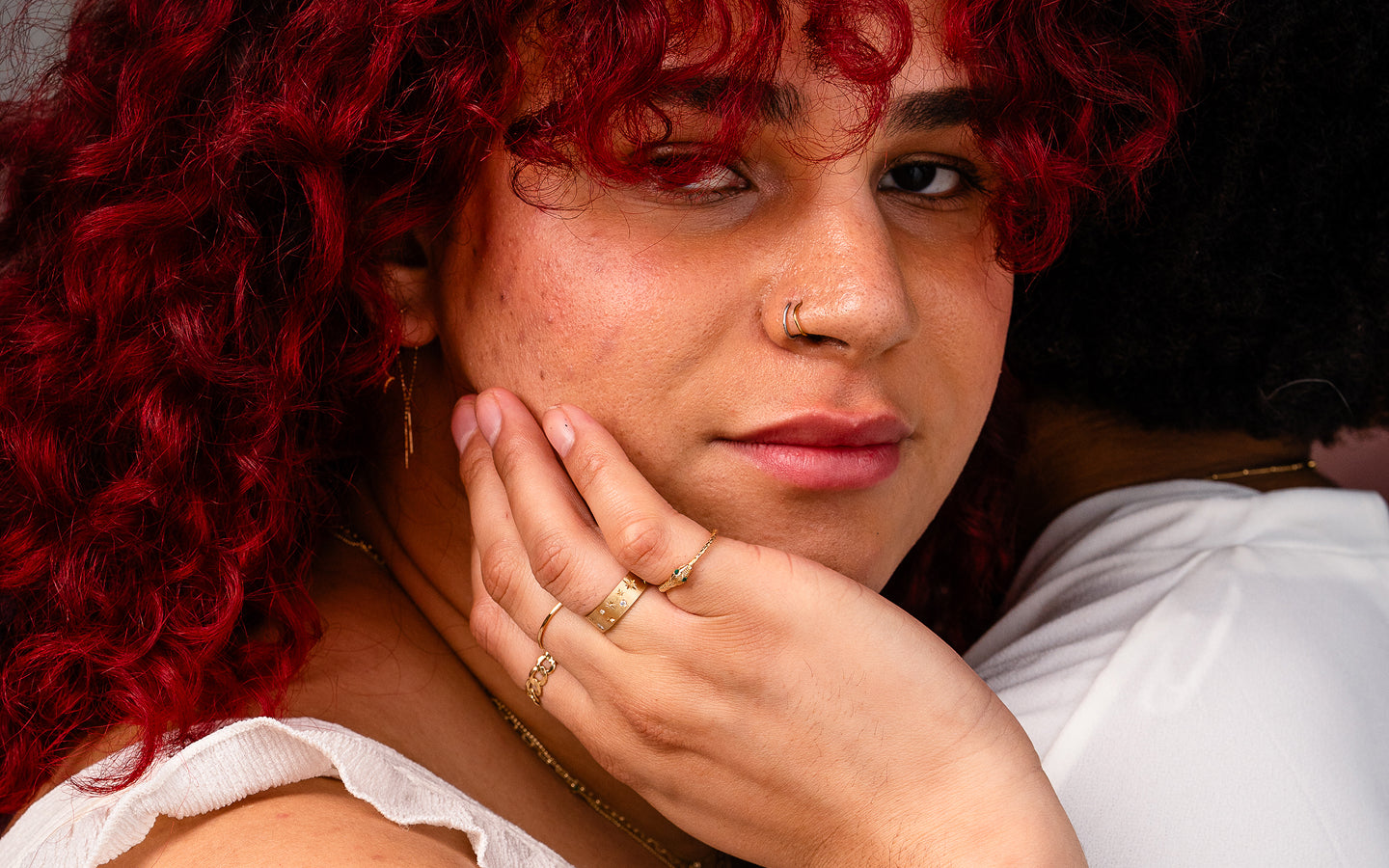 Two trans models embracing, one facing forward, caressing their face, while wearing multiple pieces of Automic Gold jewelry
