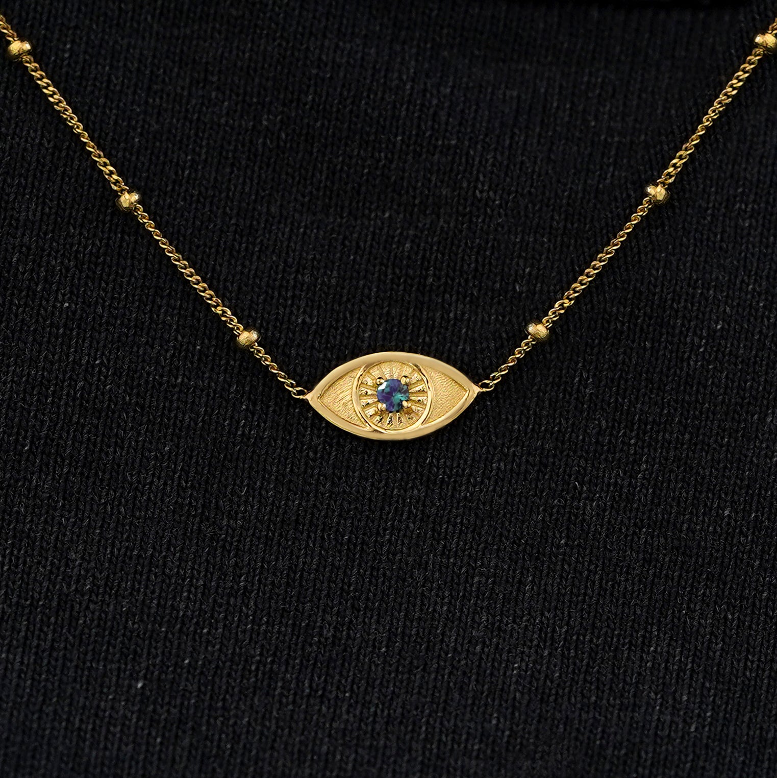 Close up view of a solid 14k yellow gold alexandrite evil eye necklace against a model's black shirt