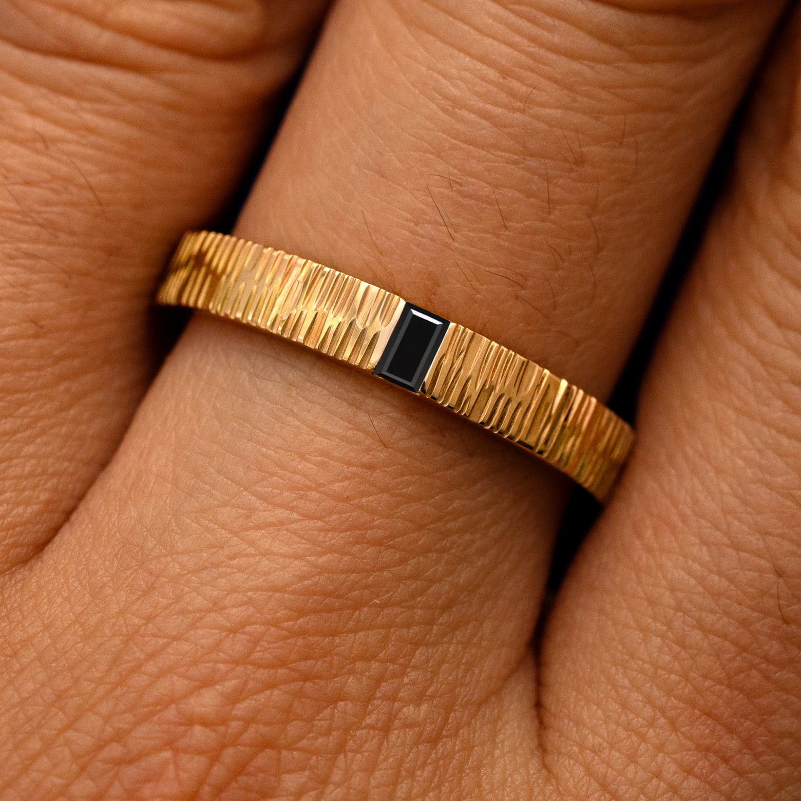 Close up view of a model's fingers wearing a 14k yellow gold Baguette Black Diamond Wood Band