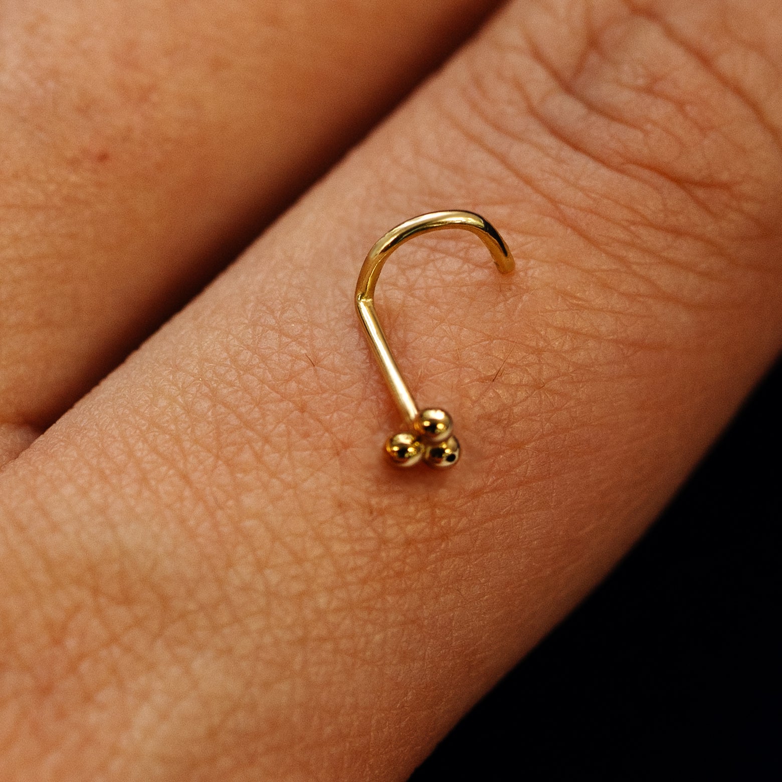 Close up view of a single 14k solid gold Ball Trio Nose Stud with a twist corkscrew backing resting on the back on a model's hand