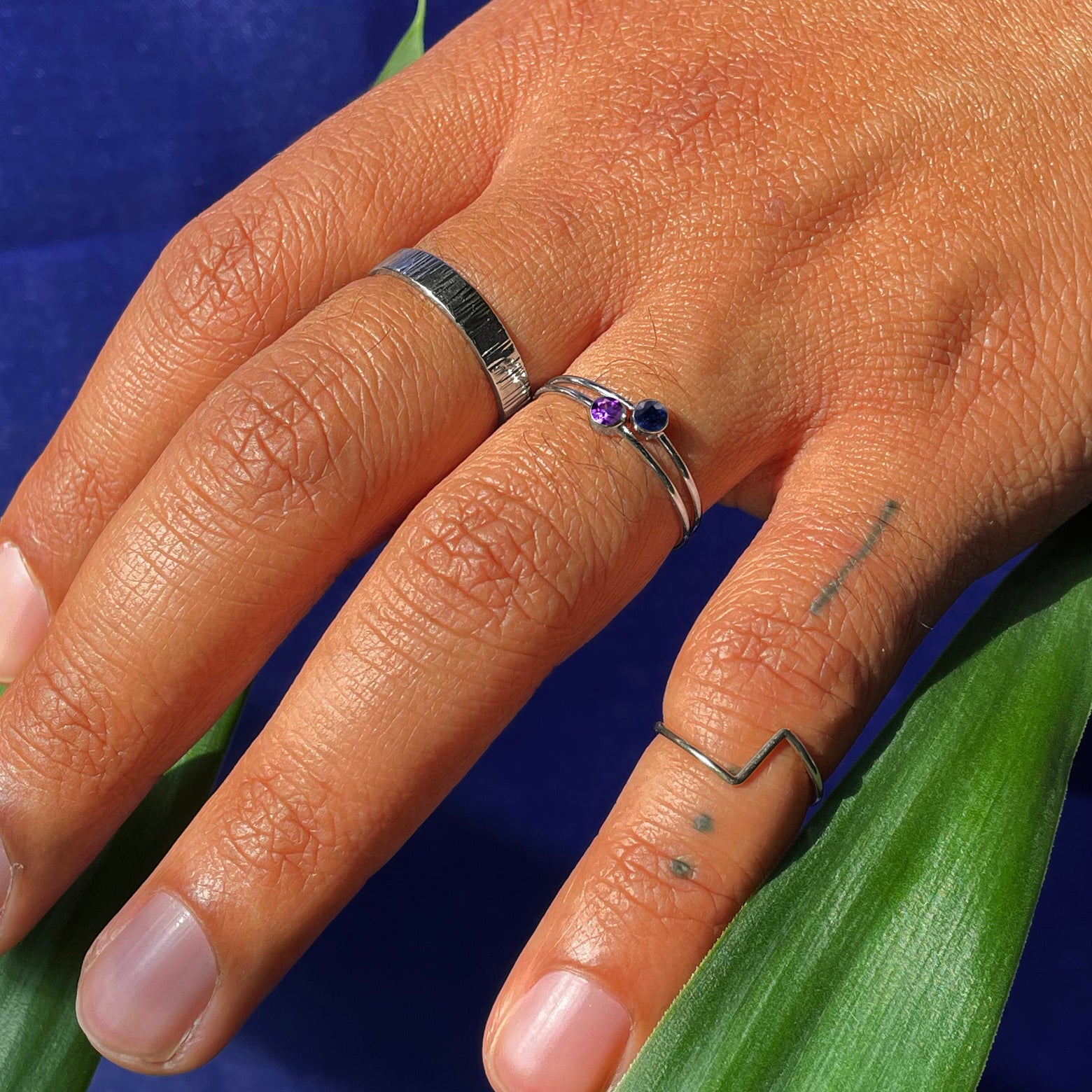 A hand touching leaves wearing an Amethyst Ring, Black Diamond Ring, Zig Zag Ring, and Industrial Wood Band all in white gold
