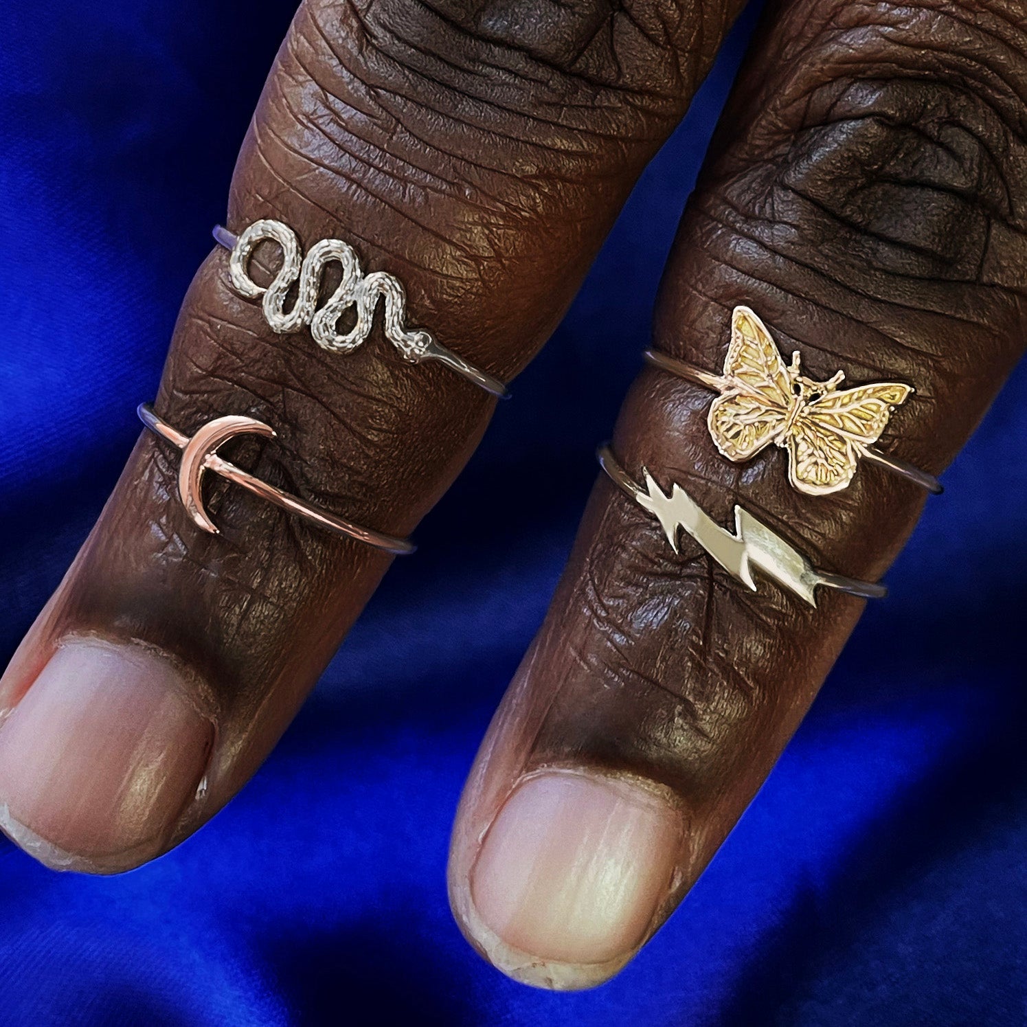 Two fingers wearing a yellow gold butterfly ring, champagne gold lightning bolt ring, white gold snake ring, and rose gold moon ring