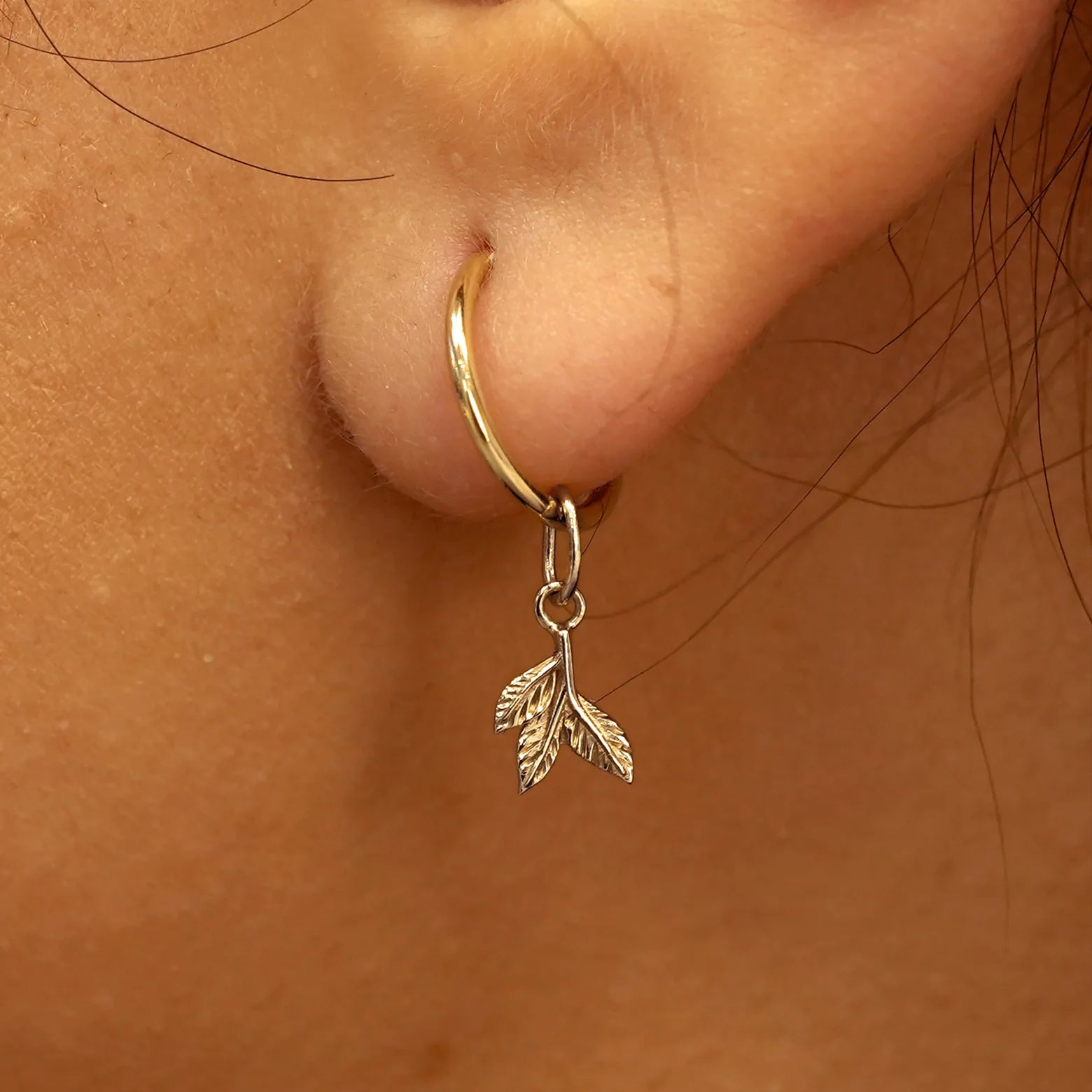 Close up view of a model's ear wearing a champagne gold 3 Leaves Charm on a Small Seamless Huggie Hoop