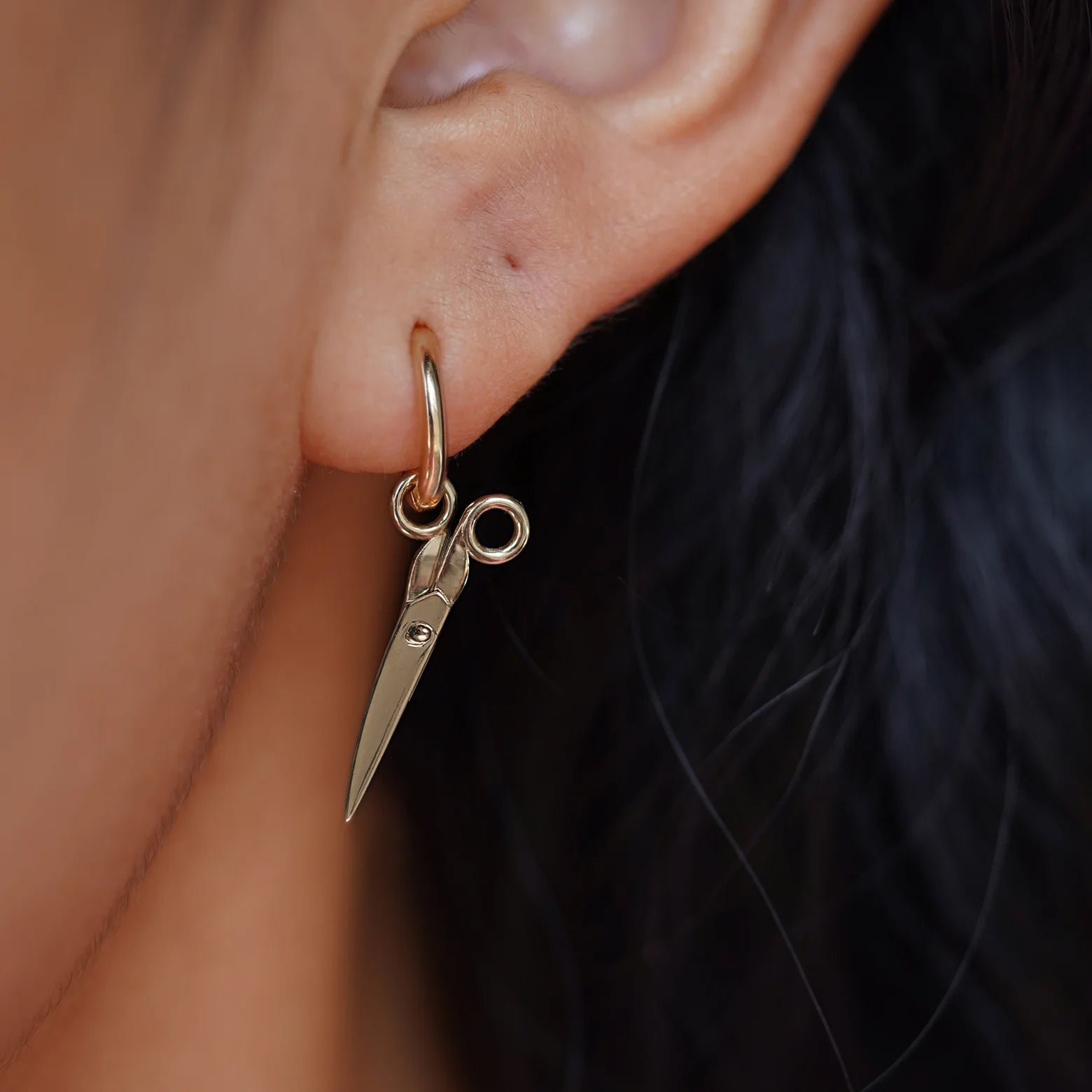 Close up view of a model's ear wearing a champagne gold Scissors Charm on a Curvy Huggie Hoop showing the scissors closed
