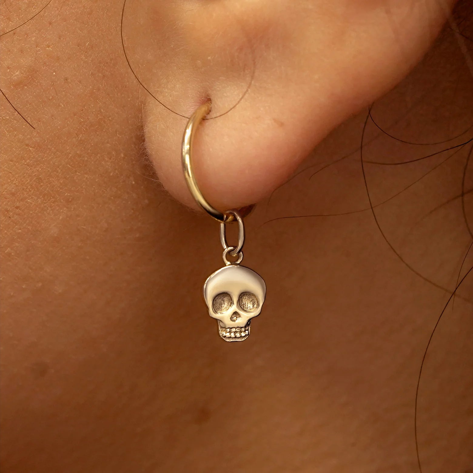Close up view of a model's ear wearing a champagne gold Skull Charm on a Small Seamless Huggie Hoop
