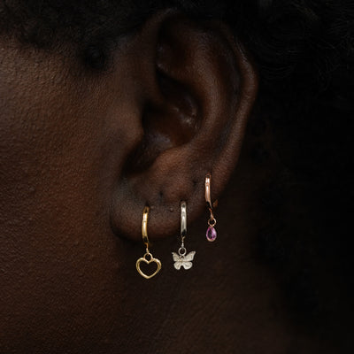 A model wearing a yellow gold heart, white gold mini butterfly, and rose gold pink sapphire dangle hoops in three piercings