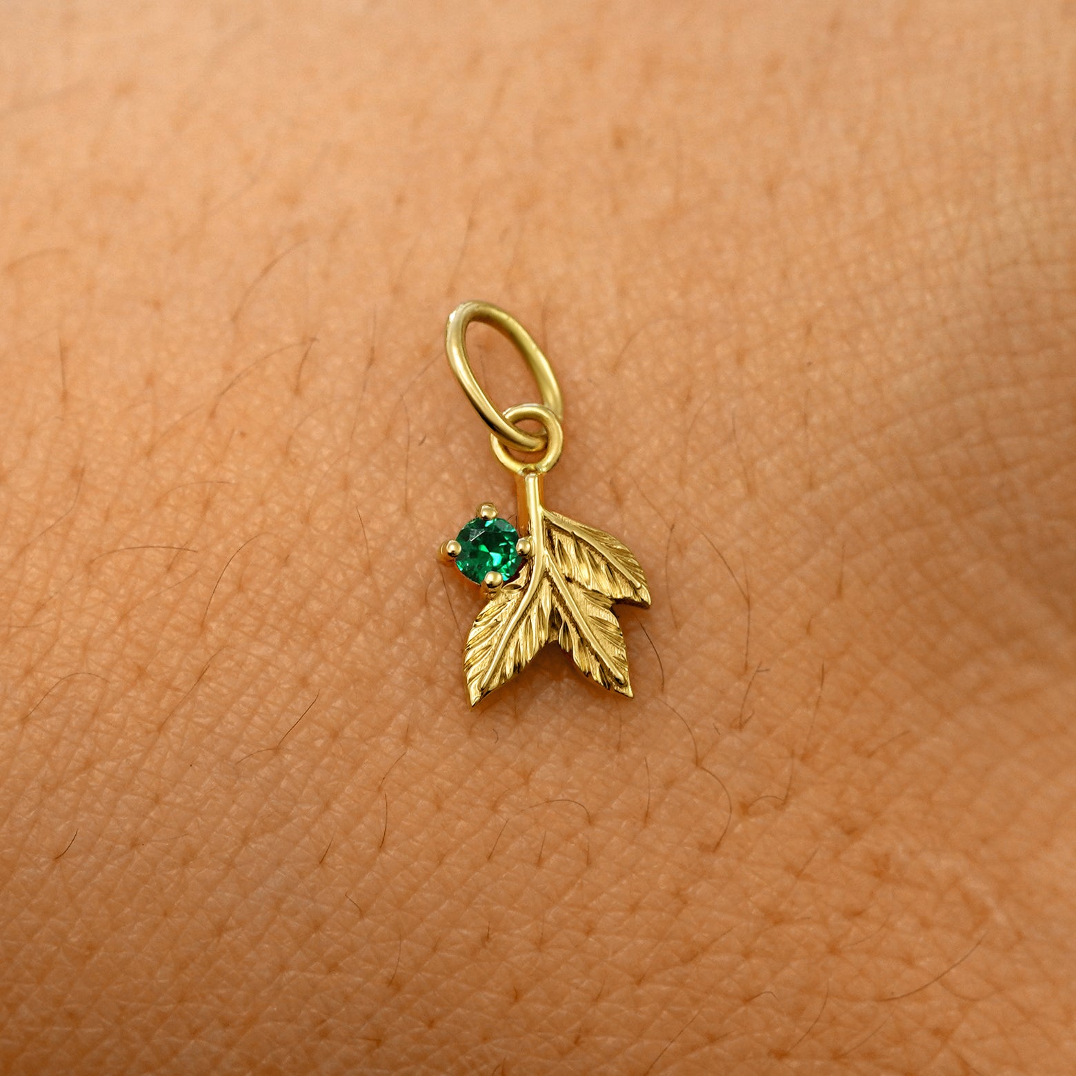 A solid 14k yellow gold emerald Gemstone 3 Leaves Charm resting on the back of a model's hand