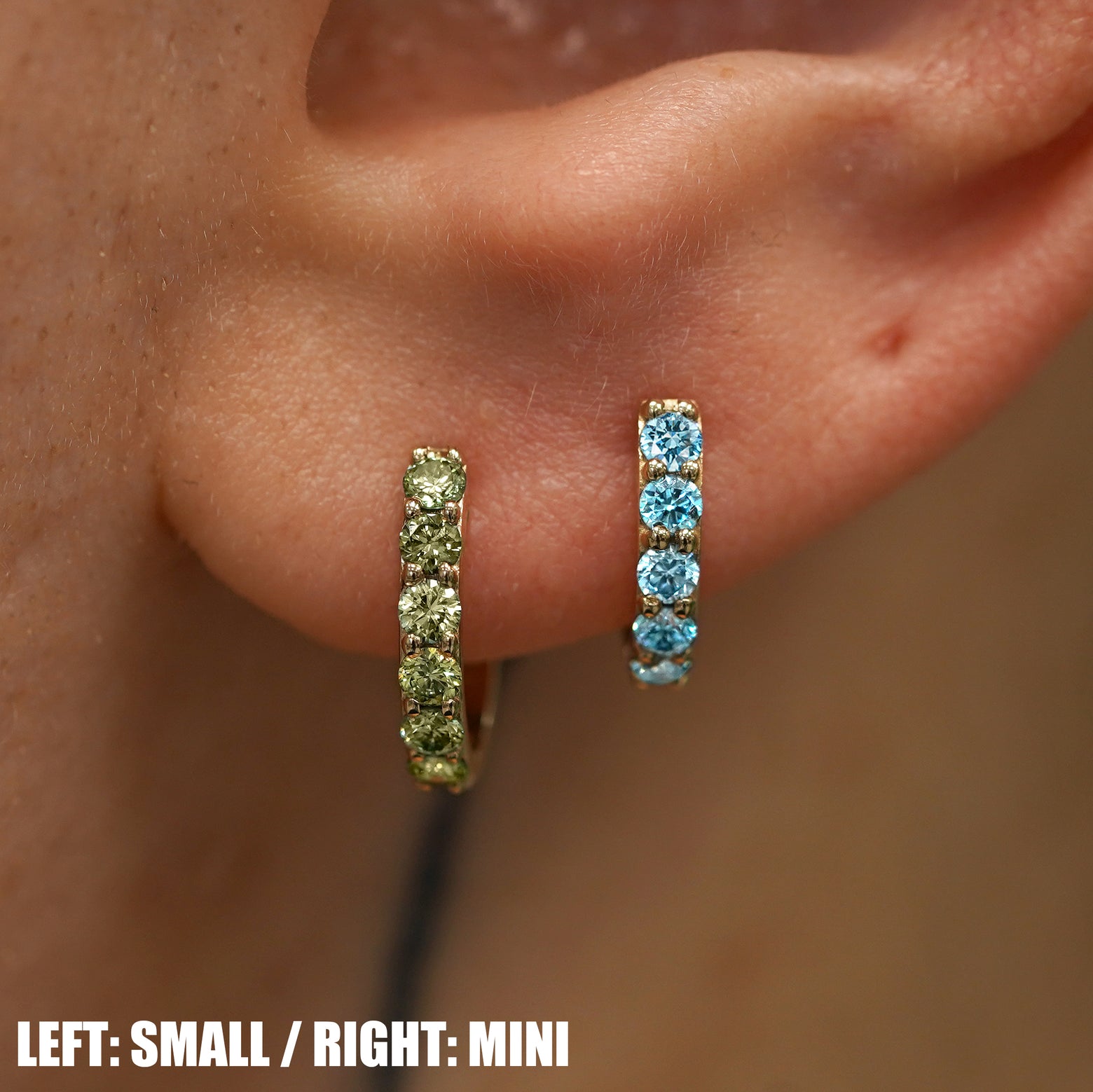 A small gemstone huggie hoop on the left a mini gemstone huggie hoop on the right on a model's finger to show size difference
