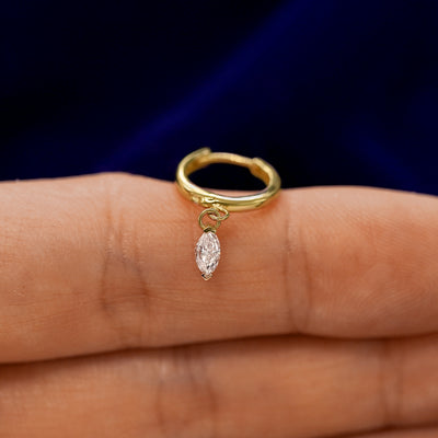 A small curvy huggie hoop earring with a dangling marquise lab diamond charm balanced on a models finger