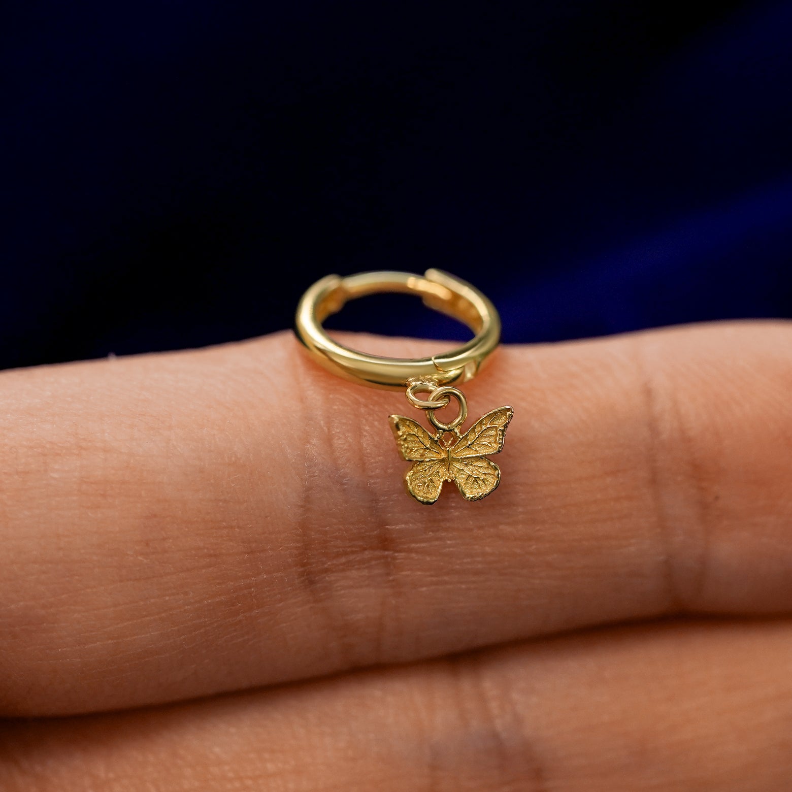 A small curvy huggie hoop earring with a dangling mini butterfly charm balanced on a models finger