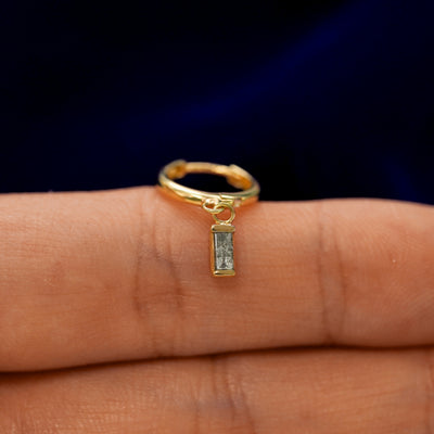 A small curvy huggie hoop earring with a dangling baguette salt and pepper charm balanced on a models finger