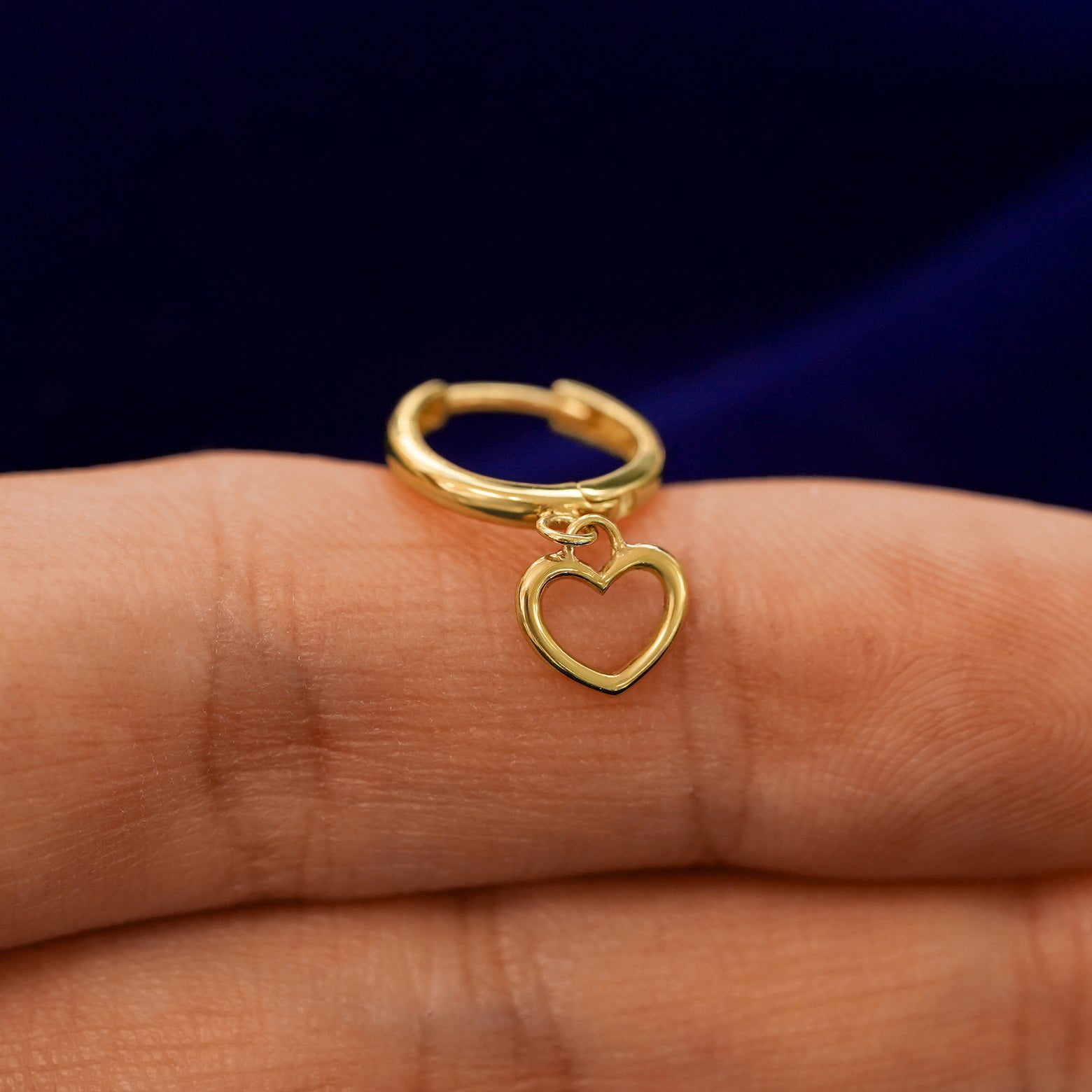 A small curvy huggie hoop earring with a dangling heart charm balanced on a models finger
