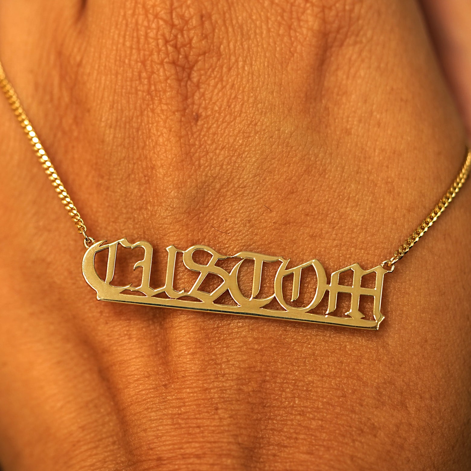 A solid yellow gold large custom nameplate necklace draped over the back of a model's hand
