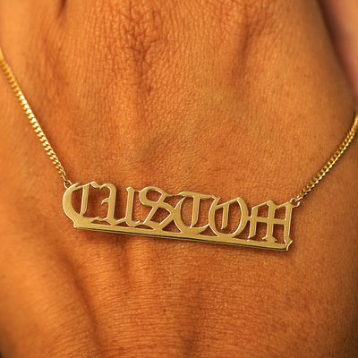 A solid yellow gold large custom nameplate necklace draped over the back of a model's hand