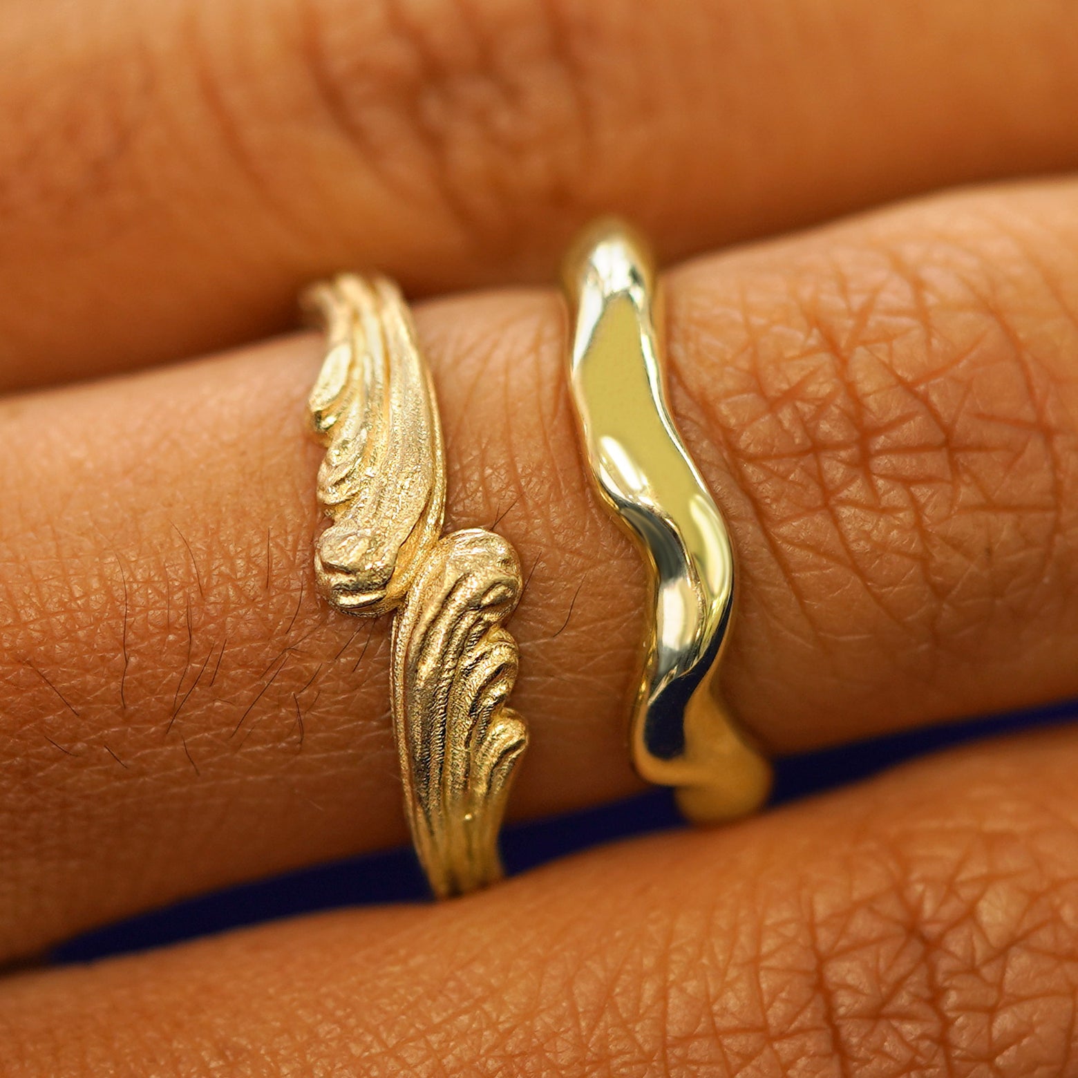 A model's hand wearing a yellow gold Liquid Band and a Crashing Waves Band stacked