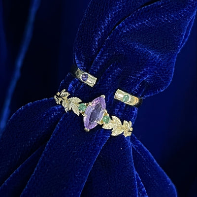 A yellow gold marquise purple sapphire leaf ring stacked with a purple and green sapphire open band on a blue cloth stand