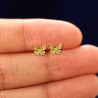 A hand holding a pair of solid yellow gold mini butterfly stud Flatback Piercings in between their fingers