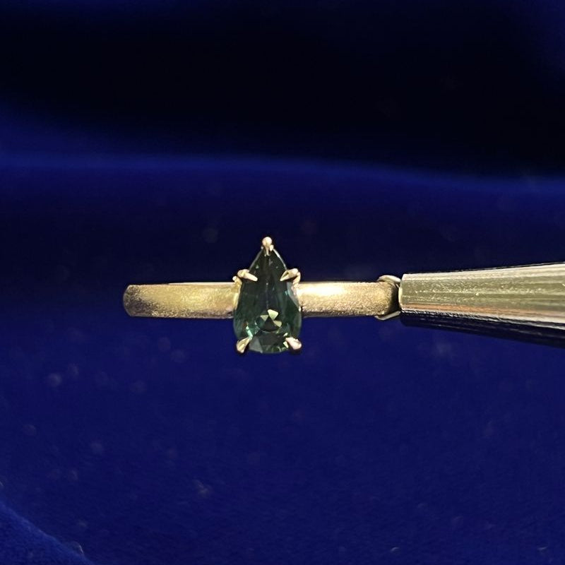 The photo displays a yellow gold matte band featuring a claw-prongs-set pear dark green sapphire.