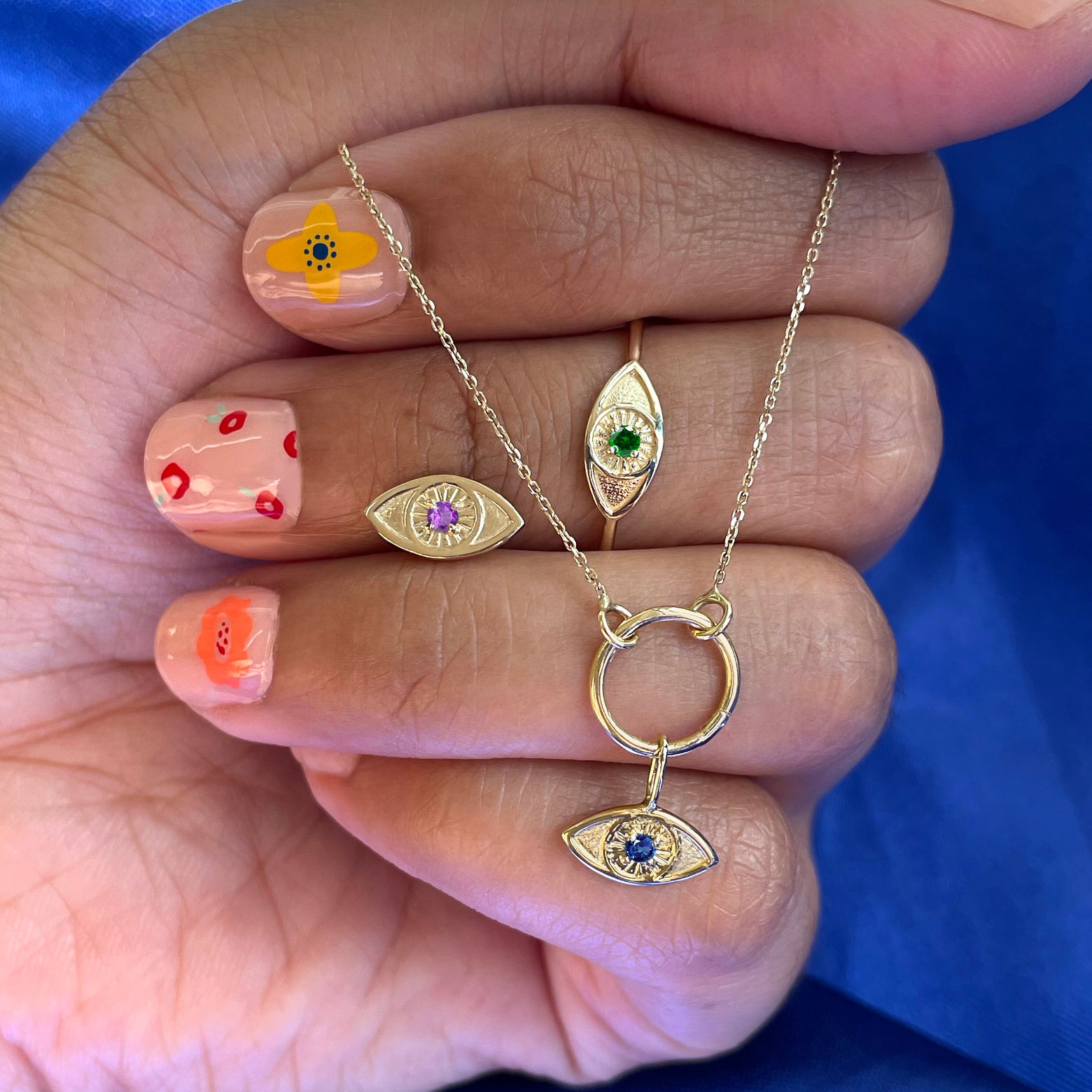 A hand wearing a Gemstone Evil Eye Ring holding an evil eye earring and a lockless cable choker with an evil eye charm 