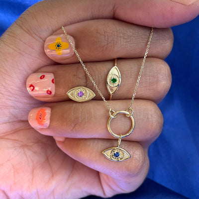 A hand wearing a Gemstone Evil Eye Ring holding an evil eye earring and a lockless cable choker with an evil eye charm 