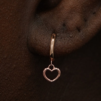 Close up view of a model's ear wearing a rose gold Heart Charm Huggie Hoop