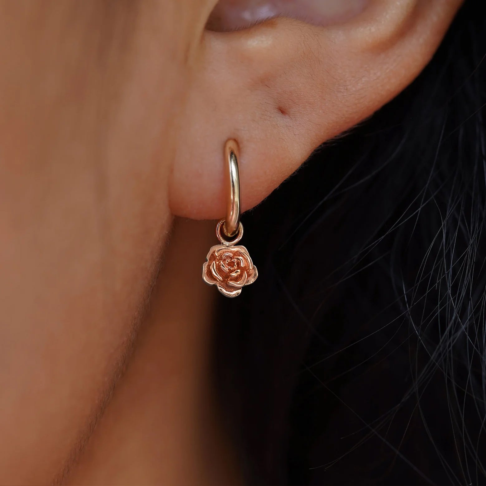 Close up view of a model's ear wearing a rose gold Rose Charm on a Small Curvy Huggie Hoop