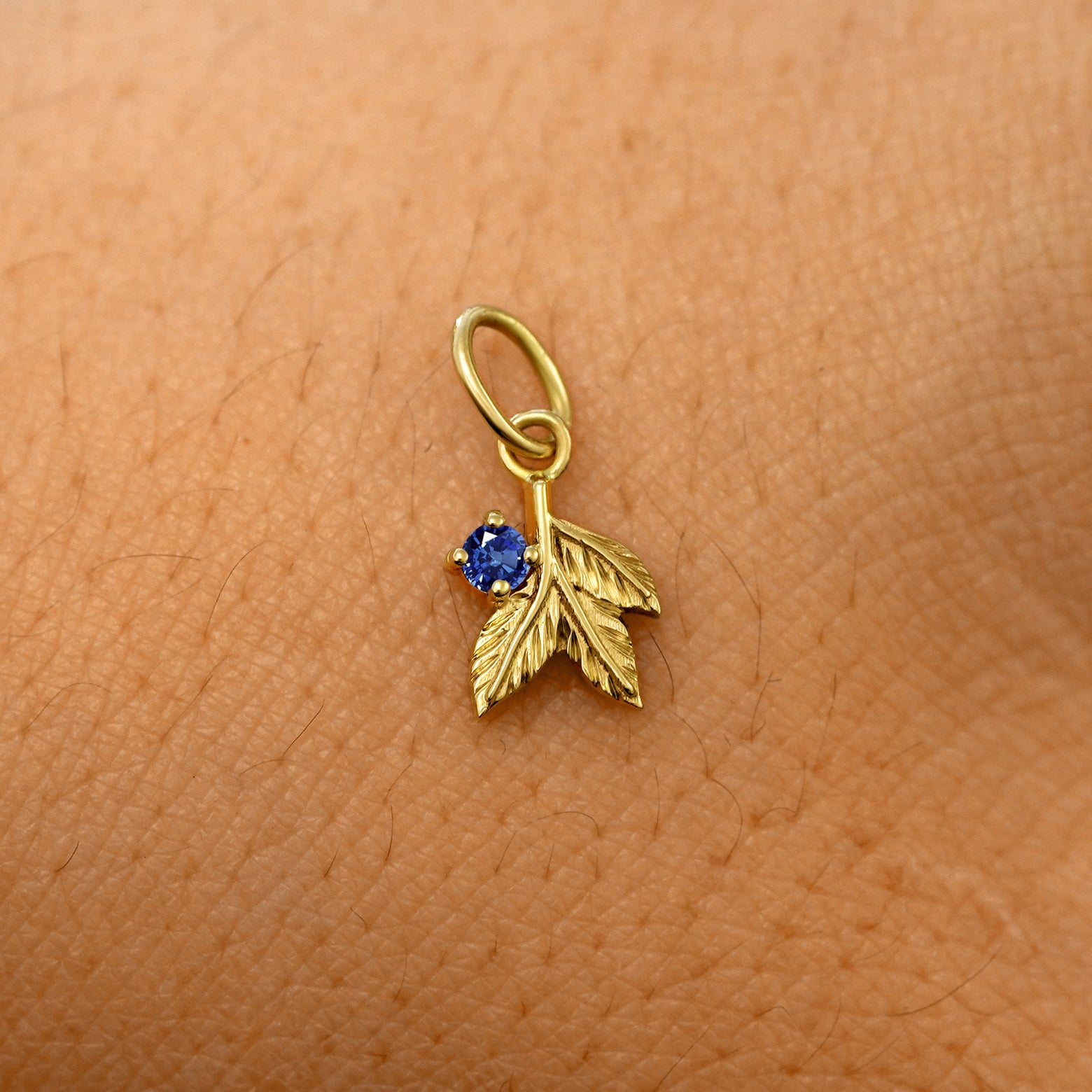 A solid 14k yellow gold blue sapphire Gemstone 3 Leaves Charm resting on the back of a model's hand