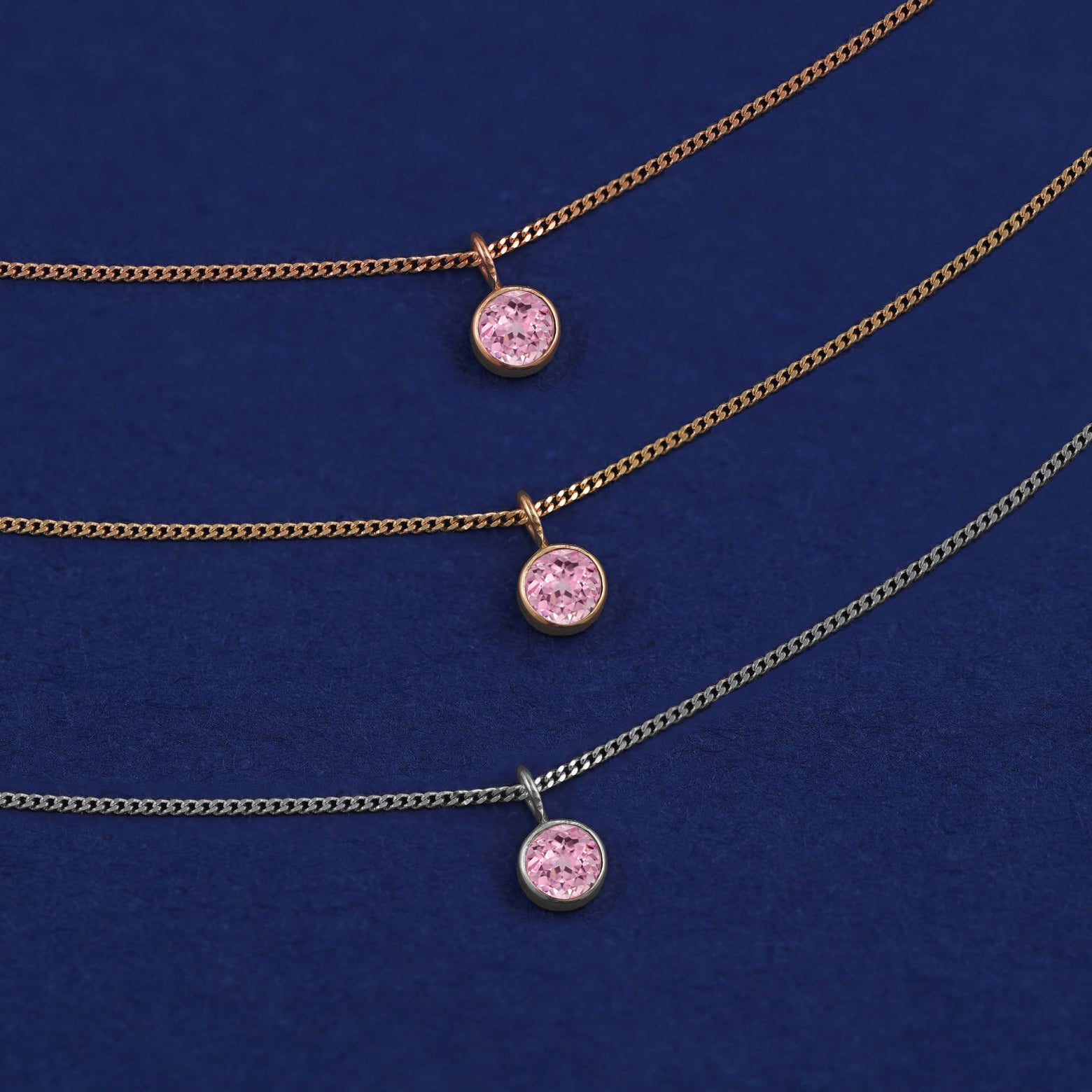 Three versions of the Pink Sapphire Necklace shown in options of yellow, white and rose gold on a dark blue background