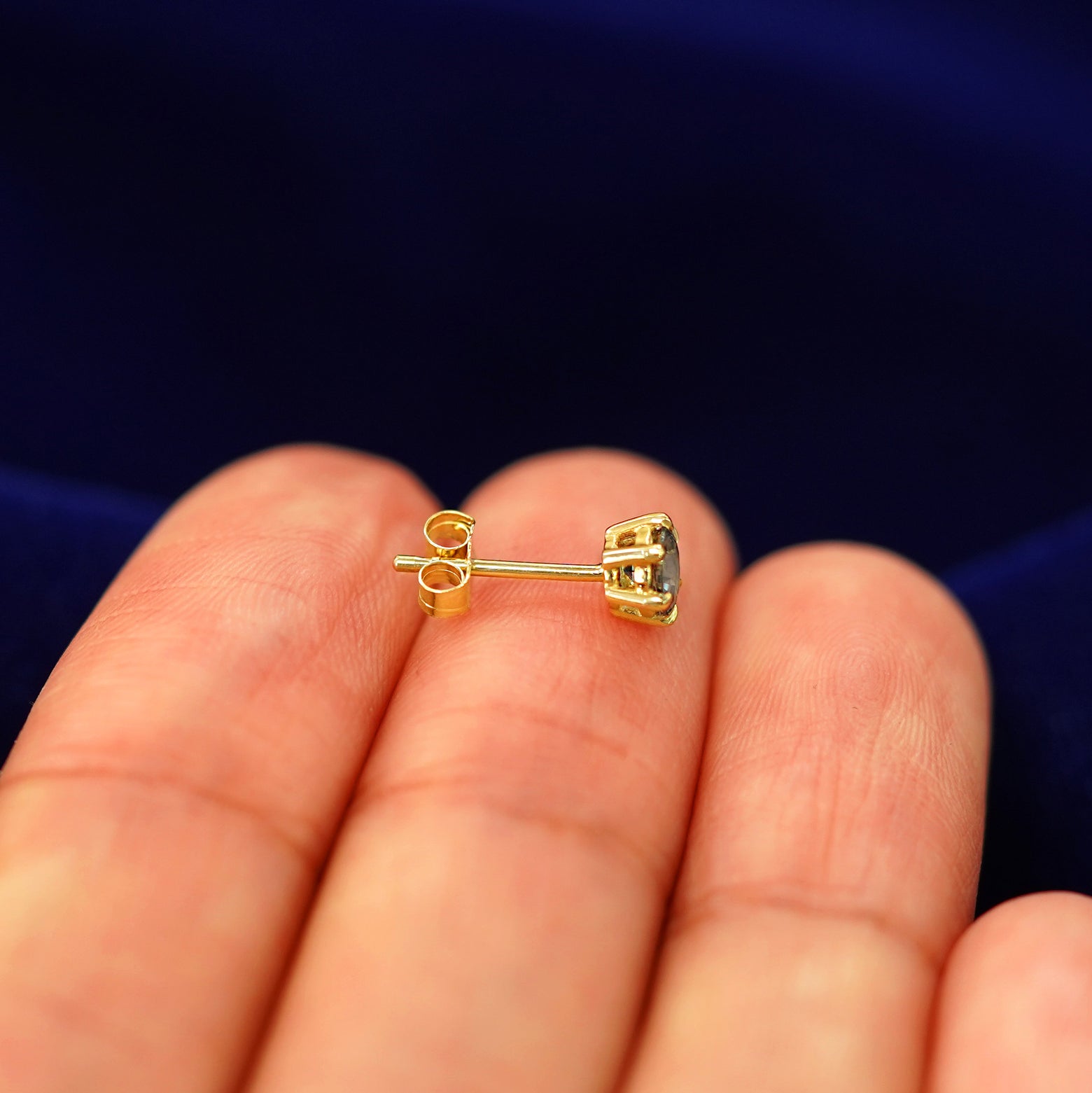 A 14k gold teal montana sapphire stud earring sitting sideways on a model's fingertips to show detail