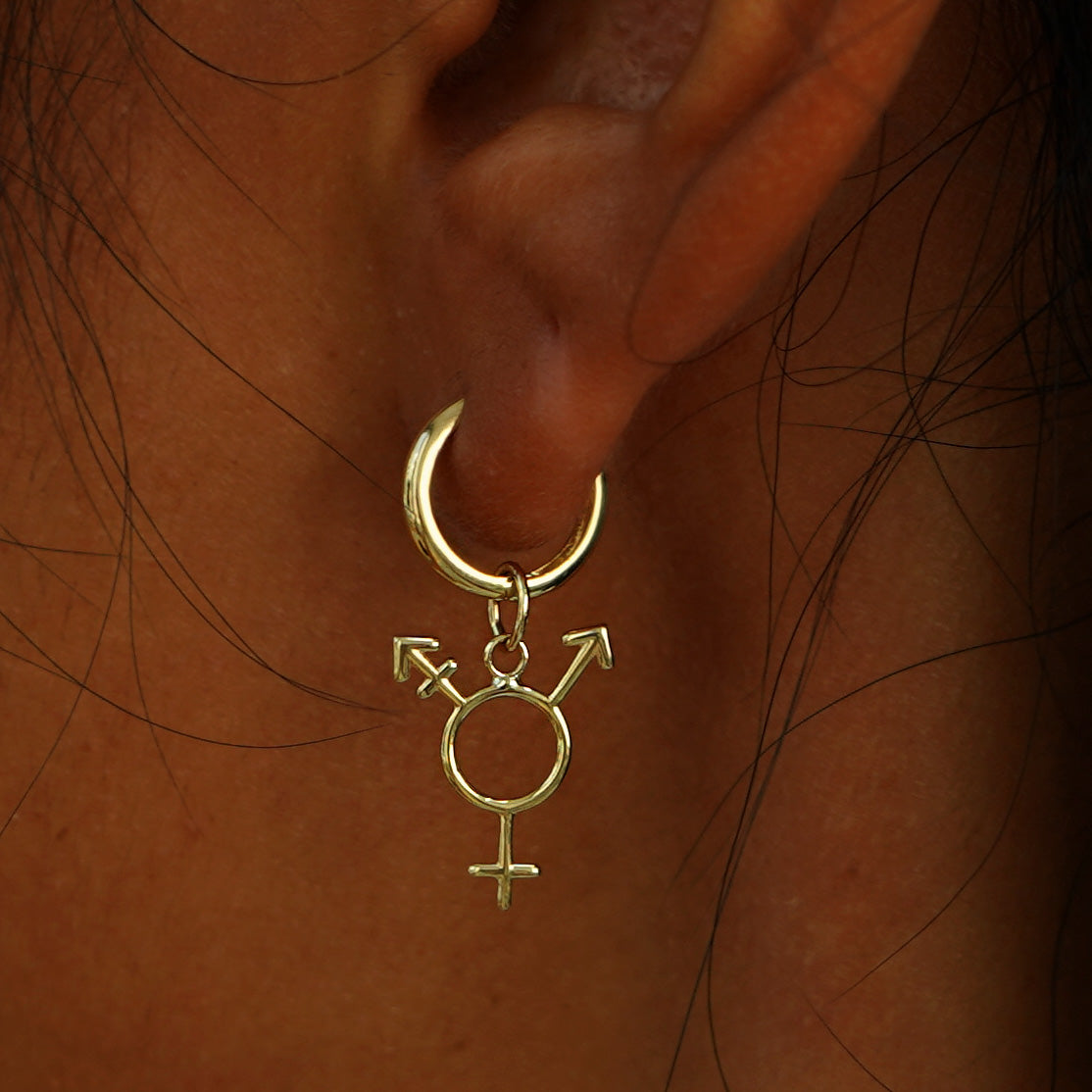 Close up view of a model's ear wearing a yellow gold Trans Symbol Charm on a Curvy Huggie Hoop