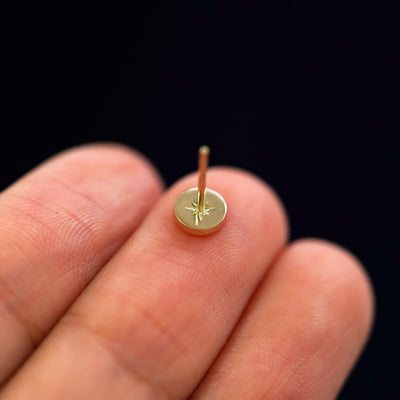 A 14k yellow gold star disk earring laying flat on a models finger with the post pointing upward