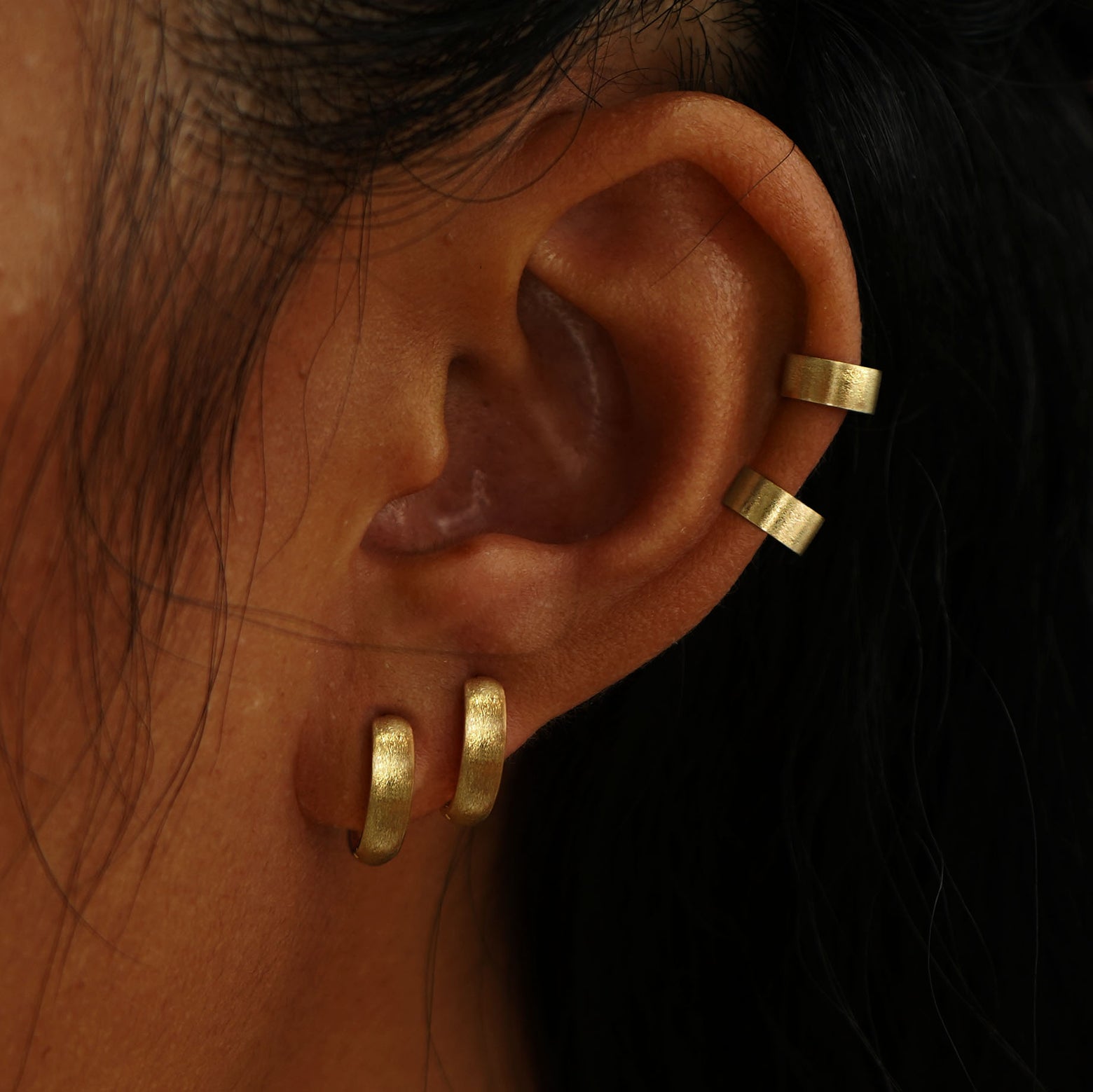 A model's ear wearing two  yellow gold Stardust Thick Cuffs and two Stardust Small Wide Huggie Hoops