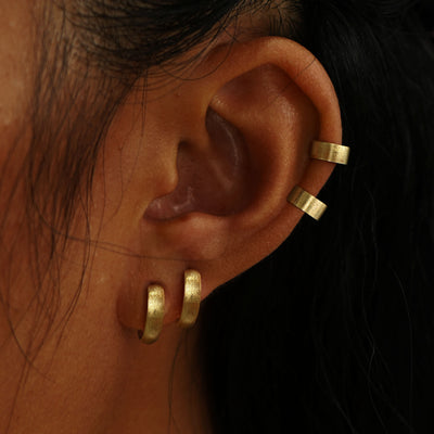 A model's ear wearing two  yellow gold Stardust Thick Cuffs and two Stardust Small Wide Huggie Hoops