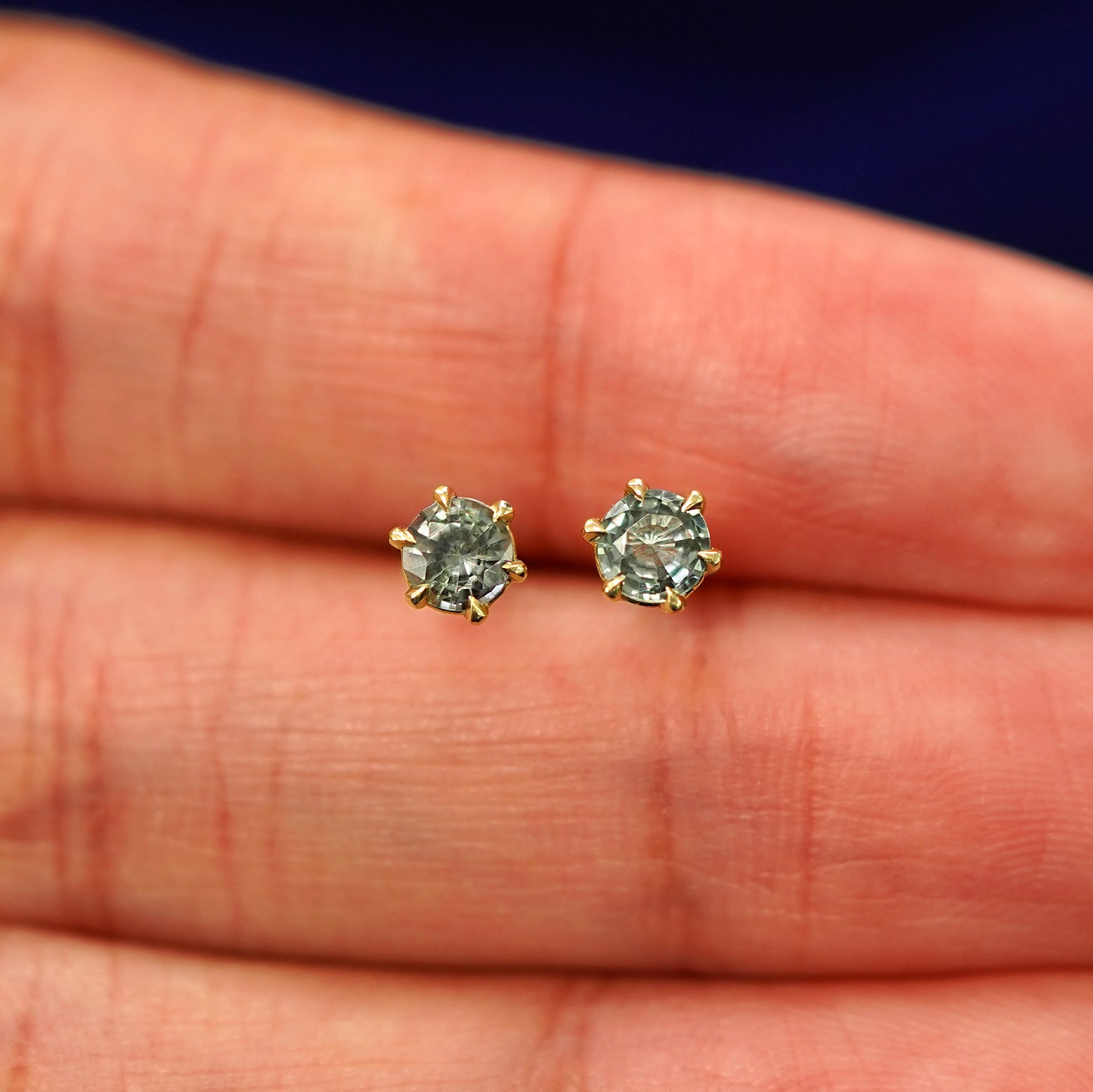 A pair of solid yellow gold clear Montana Teal Sapphire Earrings in between a model's fingers
