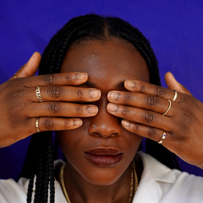 A model covering their eyes with their fingers wearing a Curb Ring, a Rainbow Band, and other Automic Gold Rings