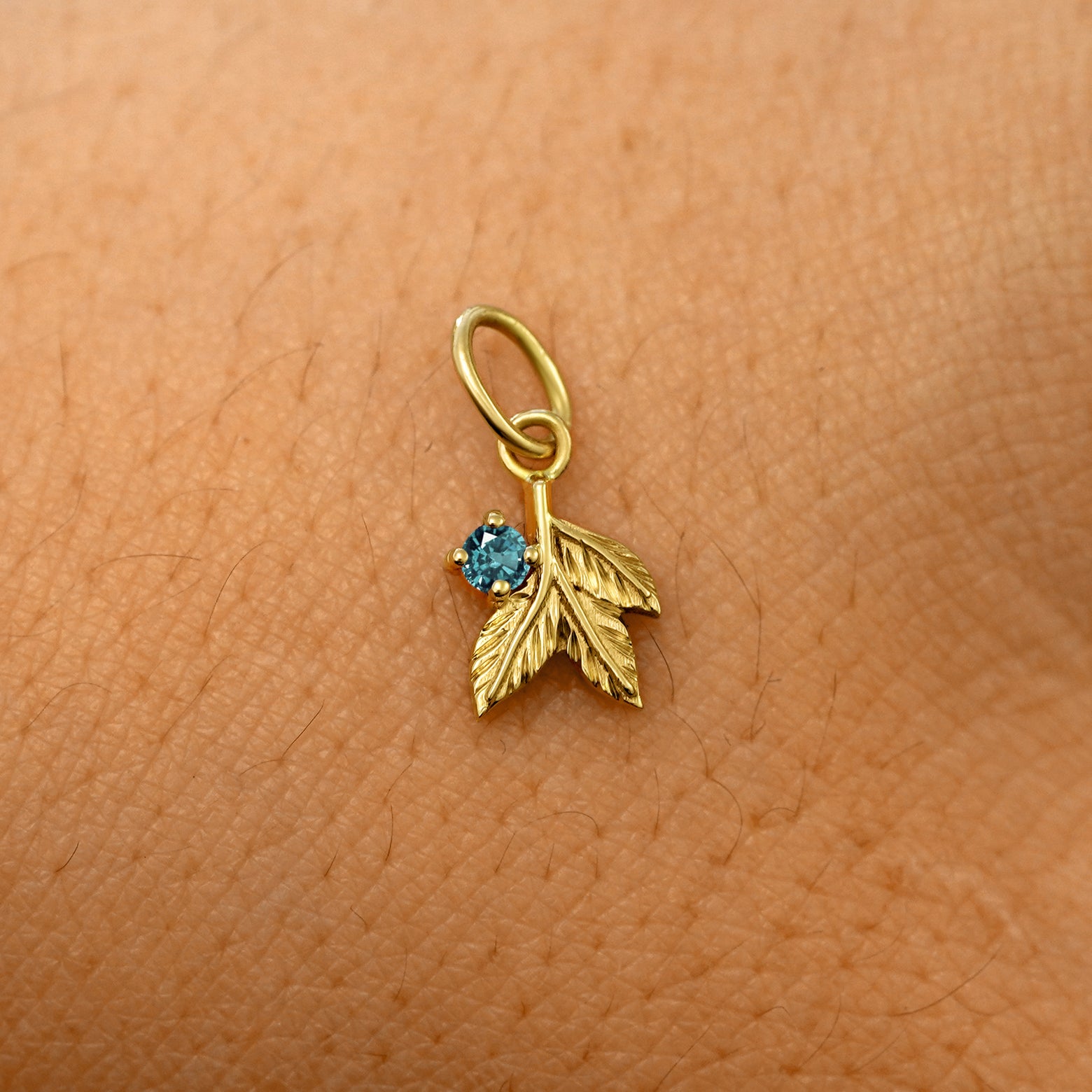 A solid 14k yellow gold blue topaz Gemstone 3 Leaves Charm resting on the back of a model's hand