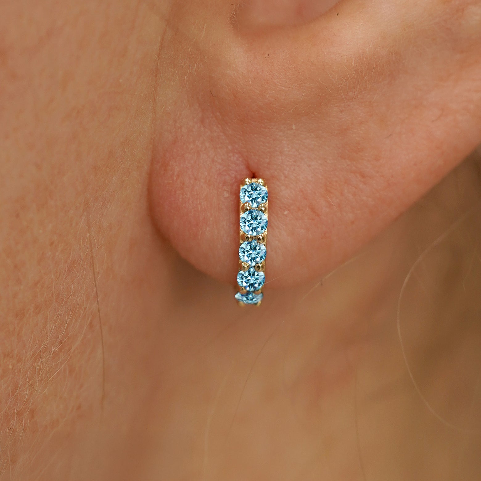 Close up view of a model's ear wearing a yellow gold Mini Light Blue Lab Diamond Huggie Hoop