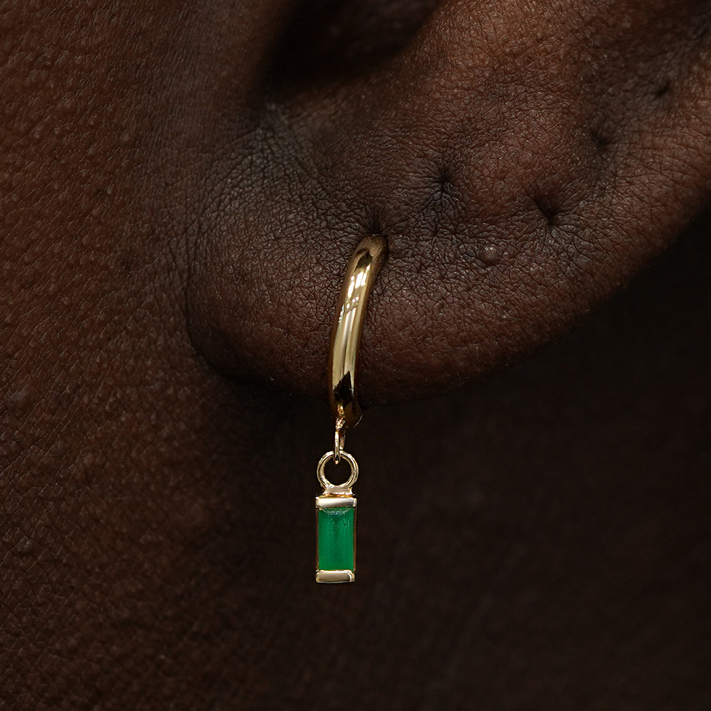 Close up view of a model's ear wearing a yellow gold Baguette Emerald Dangle Huggie Hoop