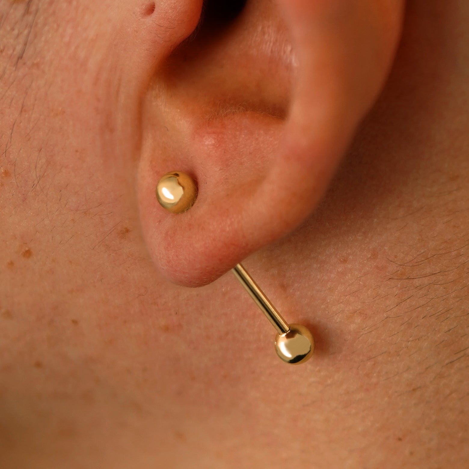 Close up view of a model's ear wearing a 14k yellow gold Large Straight Barbell through a lobe piercing