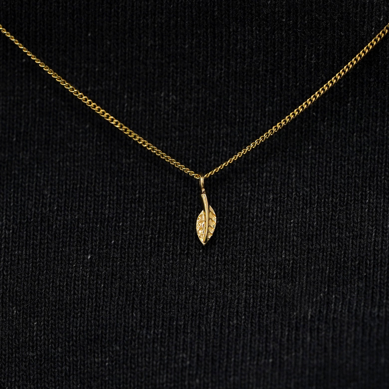 Close up view of a solid 14k yellow gold mini leaf necklace against a model's black shirt