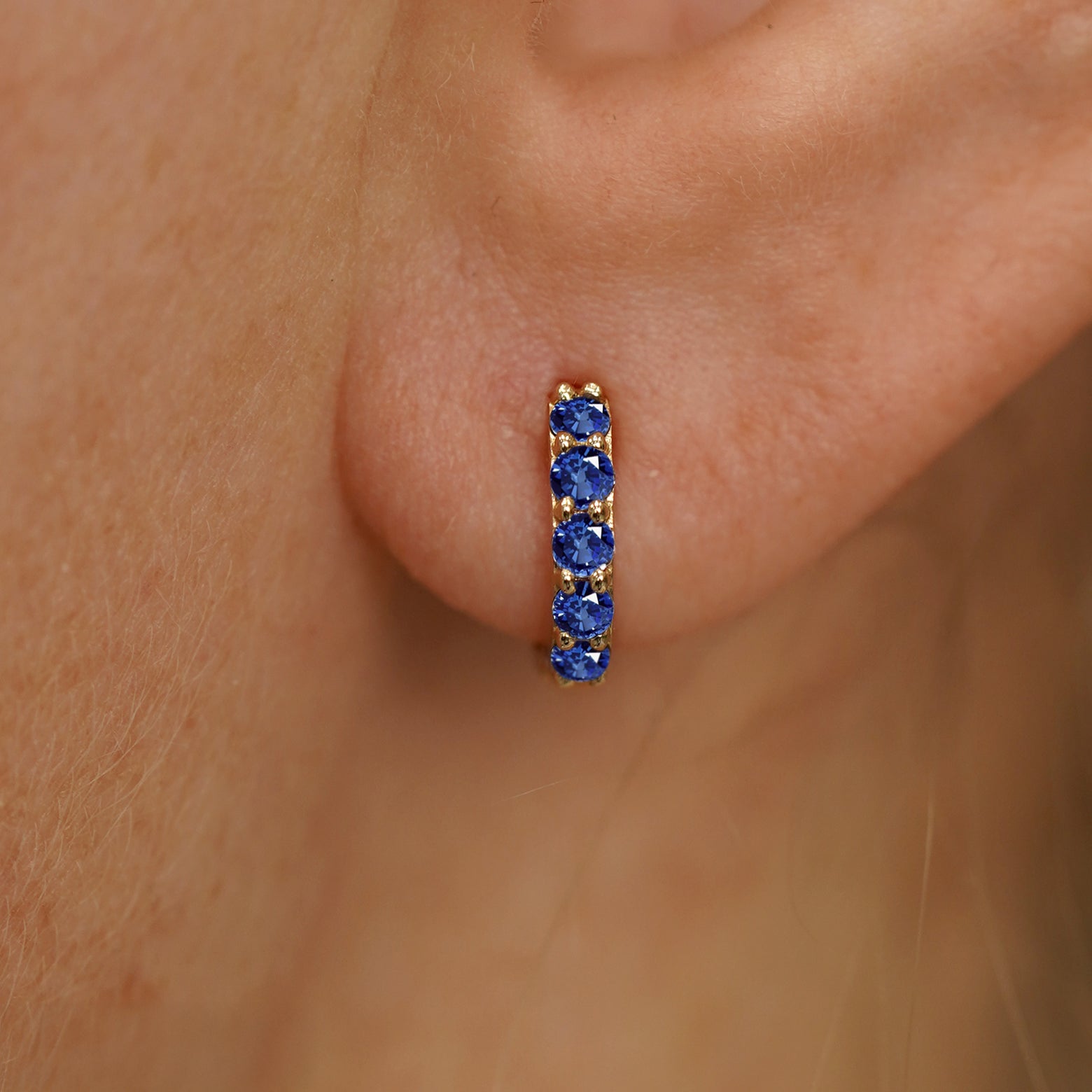 Close up view of a model's ear wearing a yellow gold Mini Blue Sapphire Huggie Hoop