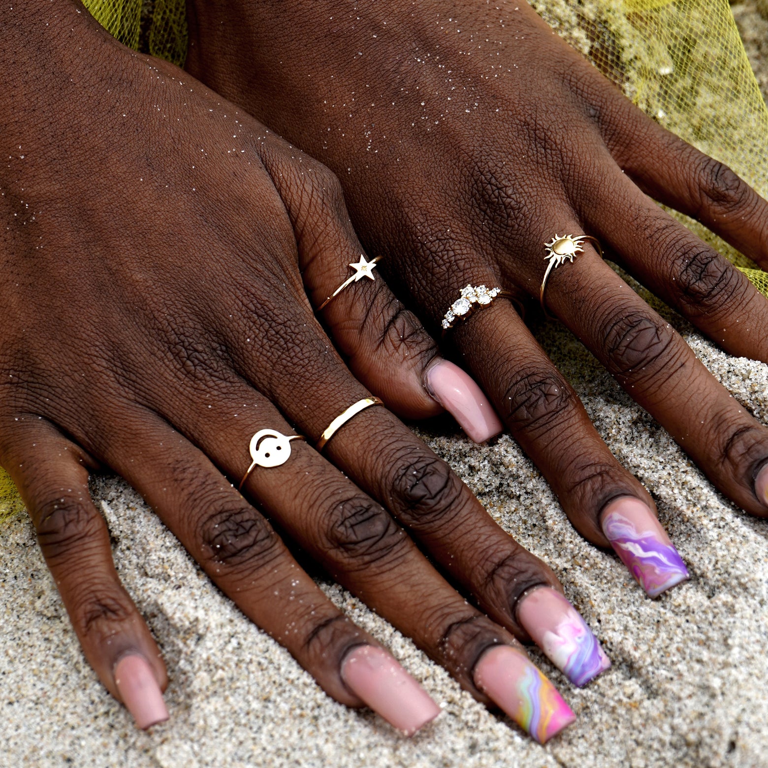A model's hands with long nails digging into sand at the beach wearing a Smiley Face Ring and other Automic Gold rings