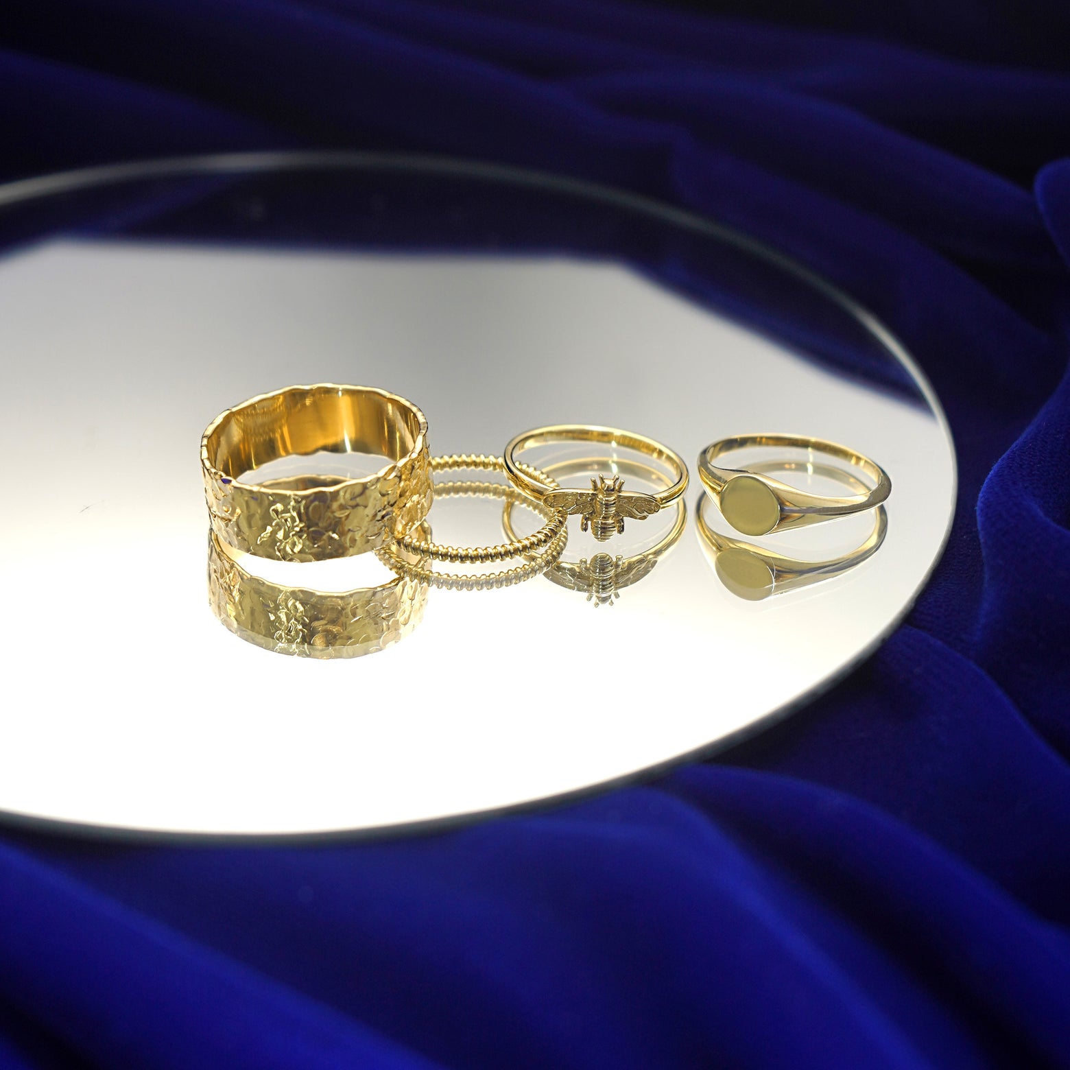 A yellow gold Industrial Hammered Band, Rope Ring, Curvy Bee Ring, and Signet Ring on a mirror