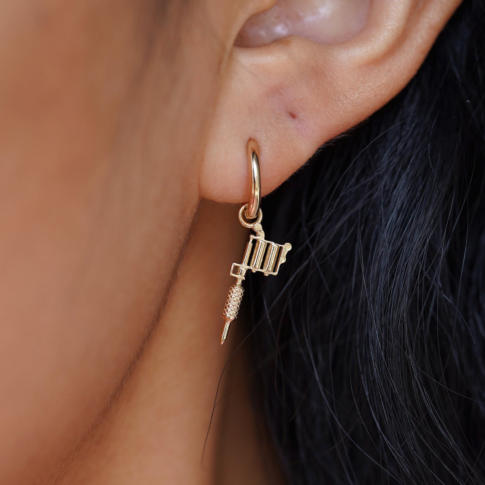 Close up view of a model's ear wearing a yellow gold Tattoo Machine Charm on a Curvy Huggie Hoop